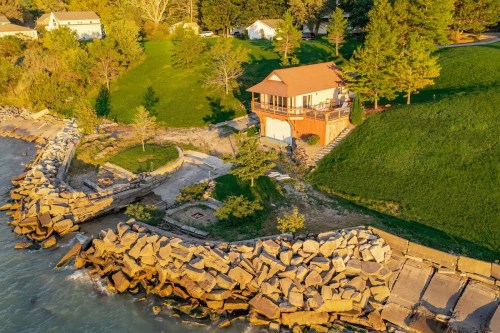 Aerial view of a house on a green hillside by the water, with a rocky shoreline and trees surrounding the area.