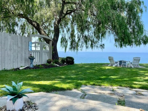 A serene lakeside view with a grassy yard, a white fence, and seating area under a willow tree.