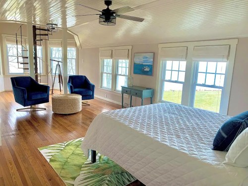 Bright, airy bedroom with a spiral staircase, two blue chairs, and large windows overlooking a grassy area.