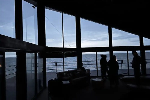 Silhouettes of people inside a modern room with large windows overlooking a calm sea and cloudy sky.