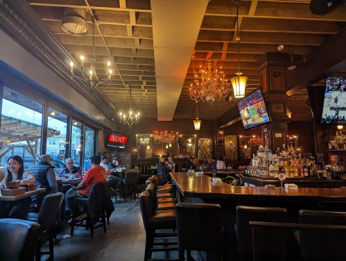 A cozy bar interior with chandeliers, a long counter, and patrons enjoying drinks and watching sports on TV.