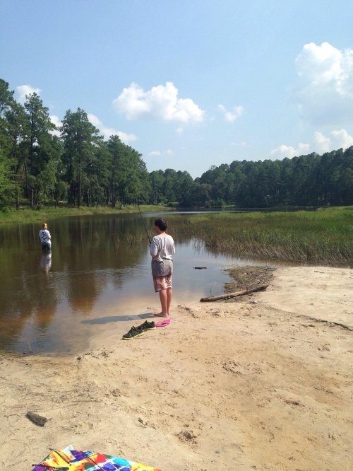 A child fishing on the shore of a calm lake, with another person wading in the water and trees in the background.