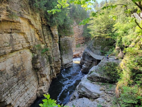 A scenic view of a rocky canyon with a river flowing through, surrounded by lush greenery and a walking path.
