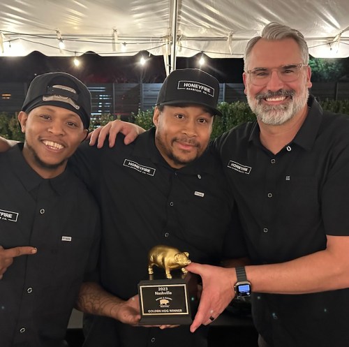Three smiling men in black shirts hold a trophy shaped like a pig under a tent at an event.