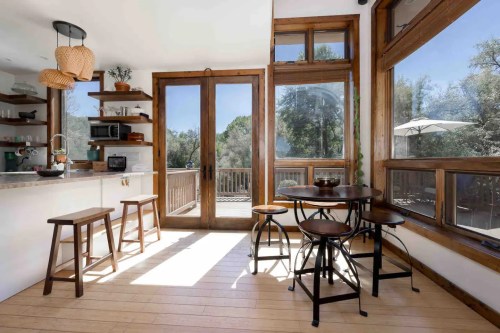 Bright, modern kitchen with wooden accents, a round table, and large windows overlooking a deck and trees.