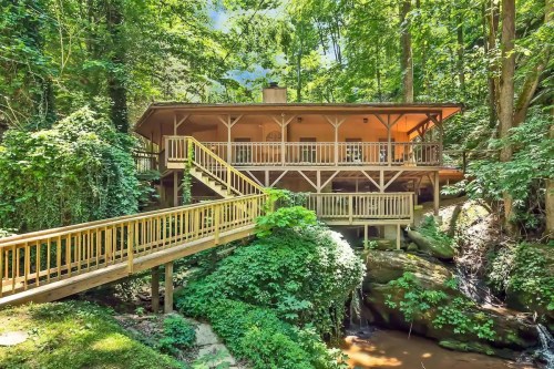 A wooden house nestled in a lush green forest, with a deck and walkway leading to a stream below.