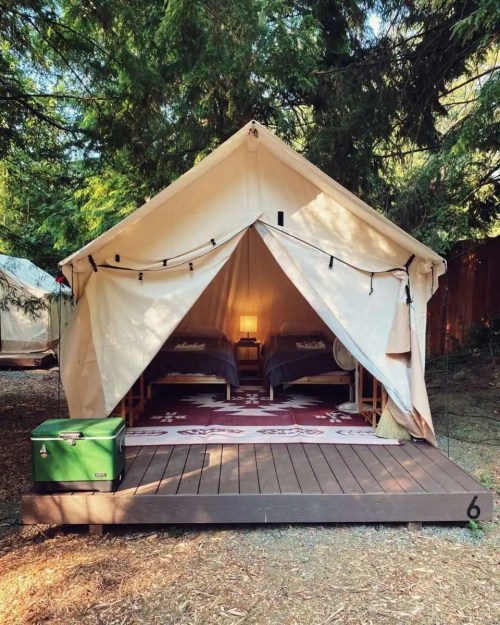 A cozy glamping tent with two beds, a rug, and a cooler outside, surrounded by trees.