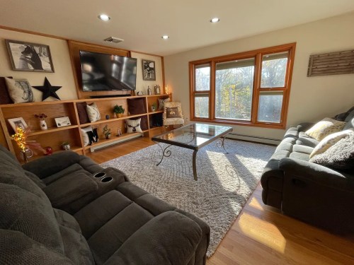 Cozy living room with a large window, gray sofas, a glass coffee table, and wooden shelves decorated with photos and plants.