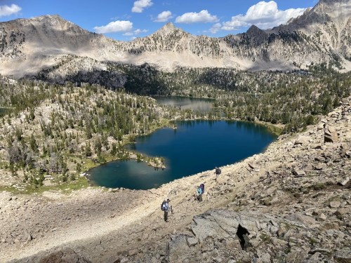 Two hikers navigate a rocky trail overlooking a serene lake surrounded by mountains and lush greenery.