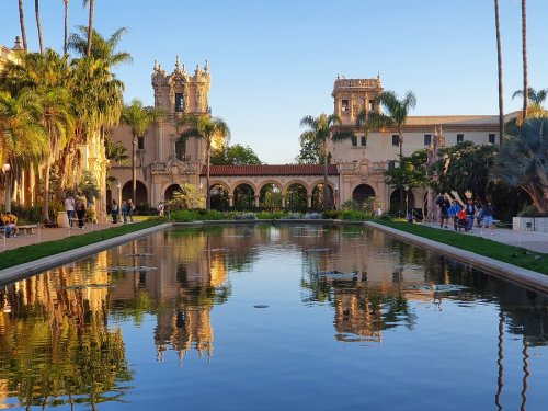 A serene park scene featuring a reflecting pond, palm trees, and historic buildings under a clear blue sky.