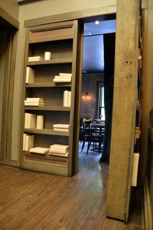 A hidden door with bookshelves leads to a dining area, featuring wooden floors and soft lighting.