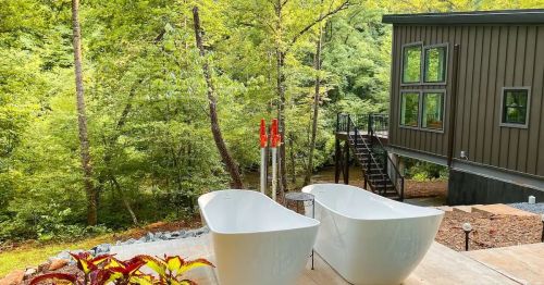 Two white bathtubs sit on a wooden deck surrounded by lush greenery and a modern house in the background.