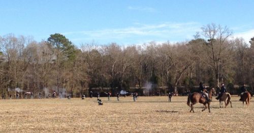 A historical reenactment scene with soldiers firing muskets in a field, horses grazing nearby, under a clear blue sky.