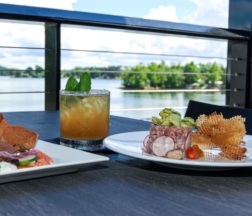 A scenic view of a lake with a cocktail and gourmet dishes on a table, surrounded by greenery and blue skies.