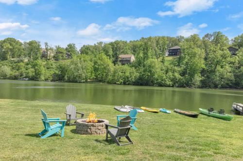 A serene lakeside scene with a fire pit, colorful chairs, and kayaks lined up on the shore, surrounded by lush greenery.