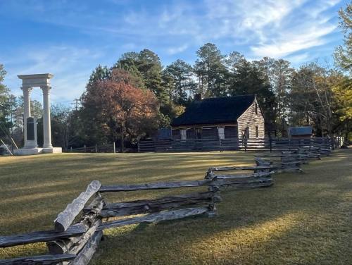 A historic site featuring a log cabin, a columned monument, and a wooden fence under a clear blue sky.