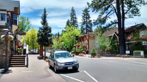 A quiet street scene with trees, shops, and a parked car under a blue sky in a small town.