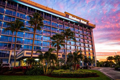 The Stadium Hotel at sunset, featuring palm trees and colorful skies reflecting on its glass facade.