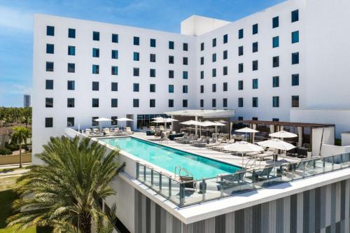Modern hotel exterior featuring a rooftop pool surrounded by lounge chairs and palm trees under a clear blue sky.