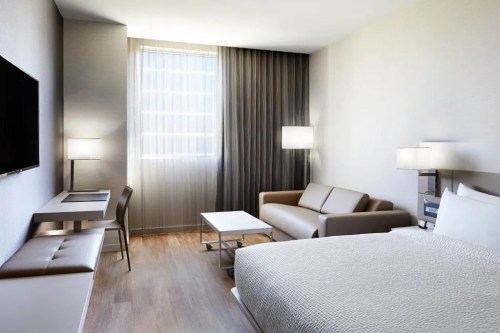 Modern hotel room featuring a bed, sofa, desk, and large window with sheer curtains.