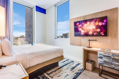 Modern hotel room featuring a bed, desk, and large windows with a view, plus a wall-mounted TV.