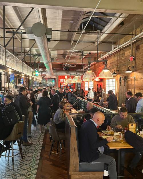 A bustling restaurant interior with patrons dining, socializing, and a bar area, featuring industrial decor and warm lighting.
