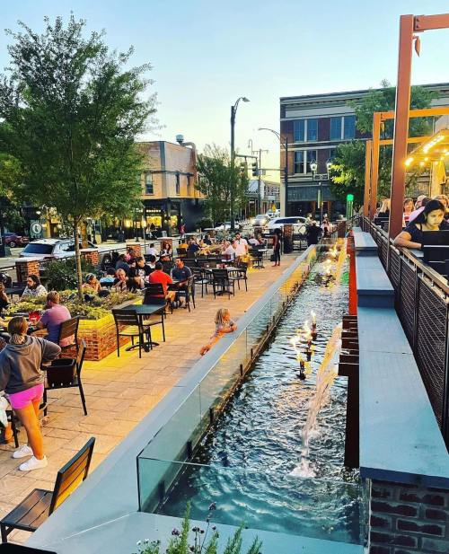 A lively outdoor dining area with people seated at tables, a water feature, and trees under a sunset sky.