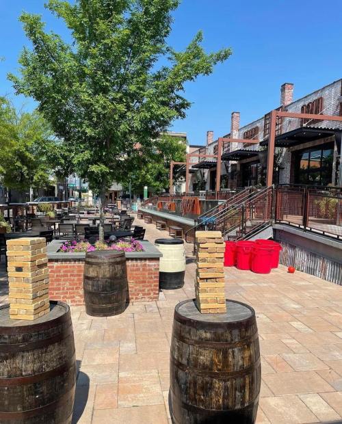 Outdoor patio with wooden barrels, stacked blocks, and a tree, surrounded by seating and buildings under a clear blue sky.