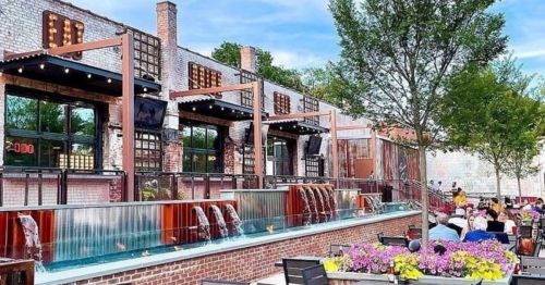 Outdoor dining area with water features, greenery, and people enjoying a sunny day at a restaurant.
