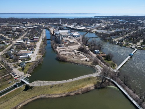Aerial view of a riverfront area with buildings, pathways, and water, surrounded by greenery and a distant lake.