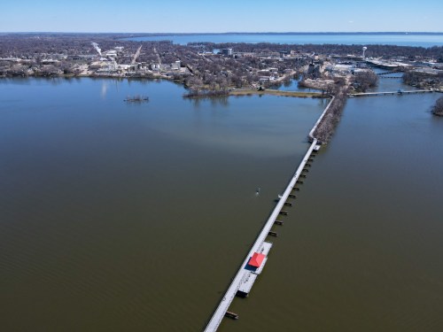 Aerial view of a river with a long pier and a red-roofed structure, surrounded by trees and a town in the background.
