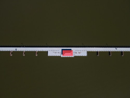 Aerial view of a long pier extending over dark water, featuring a red-roofed structure at the center.