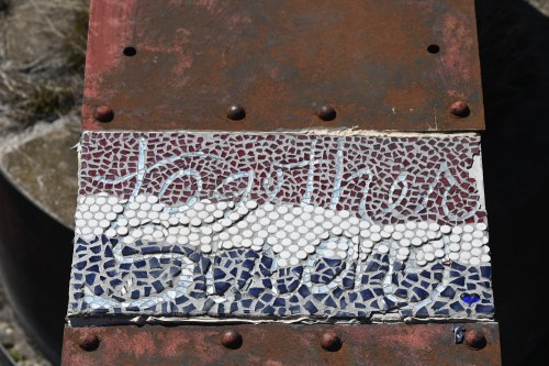 Mosaic artwork featuring abstract patterns in red, white, and blue tiles on a rusted metal surface.