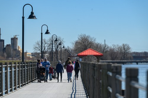 People walking along a waterfront pier with lampposts and a red-roofed gazebo on a clear day.
