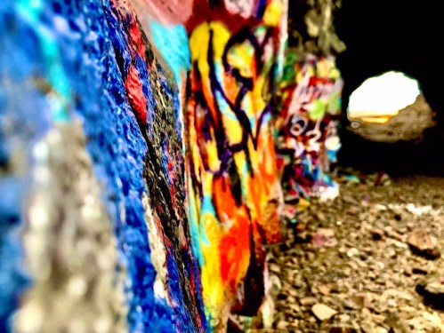 Colorful graffiti covers a rocky wall, leading into a dark tunnel with light visible at the far end.