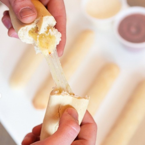 A person pulls apart a cheesy breadstick, revealing gooey cheese inside, with dipping sauces in the background.