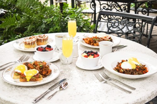 A table set with a variety of brunch dishes, including eggs Benedict, fruit, pastries, and mimosas, surrounded by greenery.