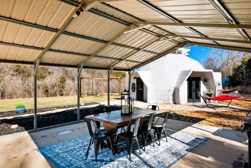 Outdoor dining area under a metal roof, featuring a table and chairs, with a view of a house and grassy yard.