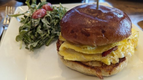 A close-up of a breakfast sandwich with eggs and bacon on a brioche bun, served with a side of arugula and cherry tomatoes.
