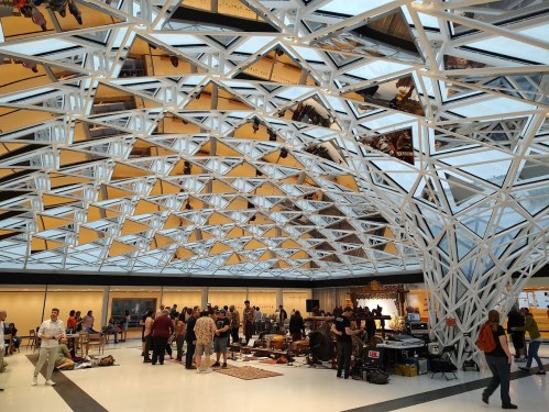 A modern indoor space with a geometric ceiling, featuring a crowd gathered around various displays and activities.