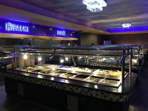 A spacious buffet with various food stations, including hibachi and sushi, illuminated by bright lights.