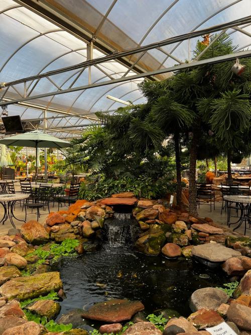 A serene indoor garden with a small pond, waterfall, and lush greenery, surrounded by tables and chairs.