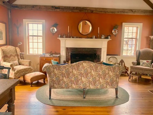 Cozy living room with a floral sofa, armchairs, a fireplace, and warm orange walls, featuring wooden floors and decorative accents.