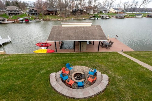 A lakeside view featuring a dock, kayaks, and a circular fire pit surrounded by colorful chairs on a grassy area.