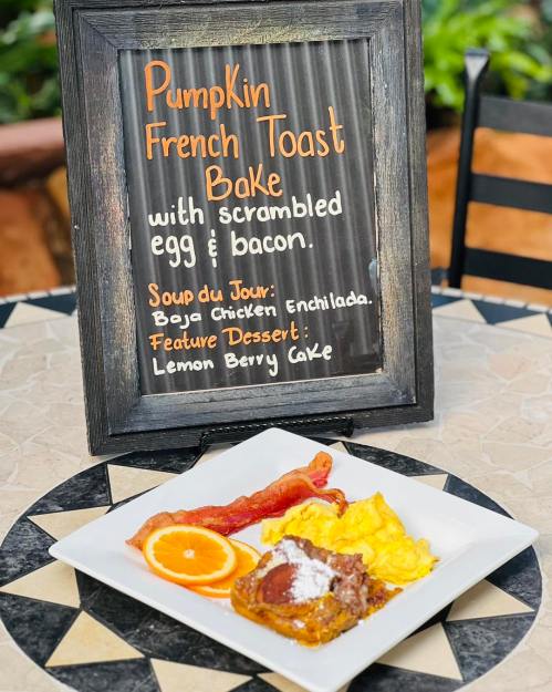 A plate of pumpkin French toast bake with scrambled eggs, bacon, and an orange slice, next to a menu sign.