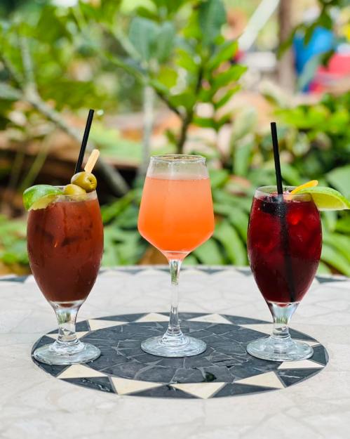 Three colorful cocktails on a patterned table, surrounded by greenery.