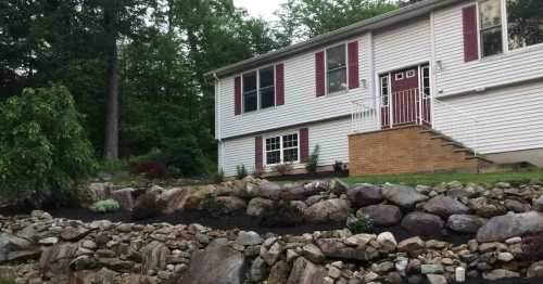 A two-story house with white siding, red shutters, and a stone garden in front, surrounded by trees.