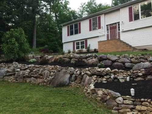 A landscaped yard featuring stone walls, flower beds, and a two-story house with red shutters and a front staircase.