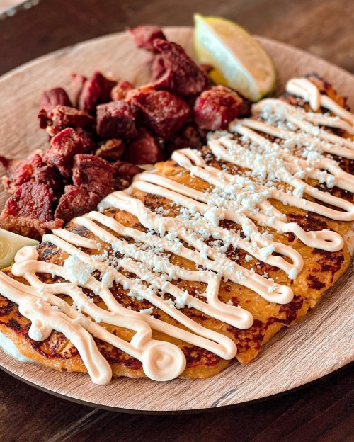 A plate featuring grilled meat, two corn cakes drizzled with sauce, and lime wedges.