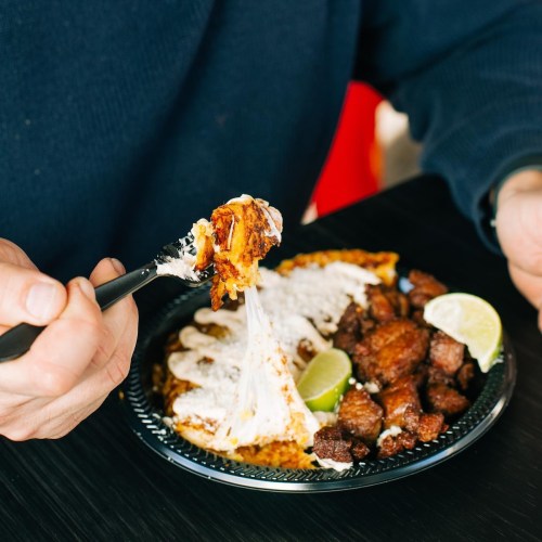 A person holds a fork with cheesy food above a plate of meat and lime wedges, ready to take a bite.